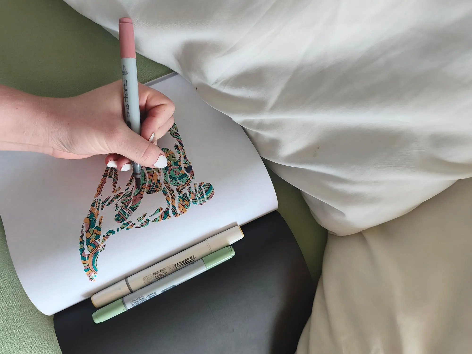 Eine Hand mit rosafarbenem Marker malt ein Surf-Motiv aus, das schon in Türkis, Orange und Grün leuchtet. Das Buch liegt auf heller Bettwäsche, zwei weitere Marker liegen quer davor.