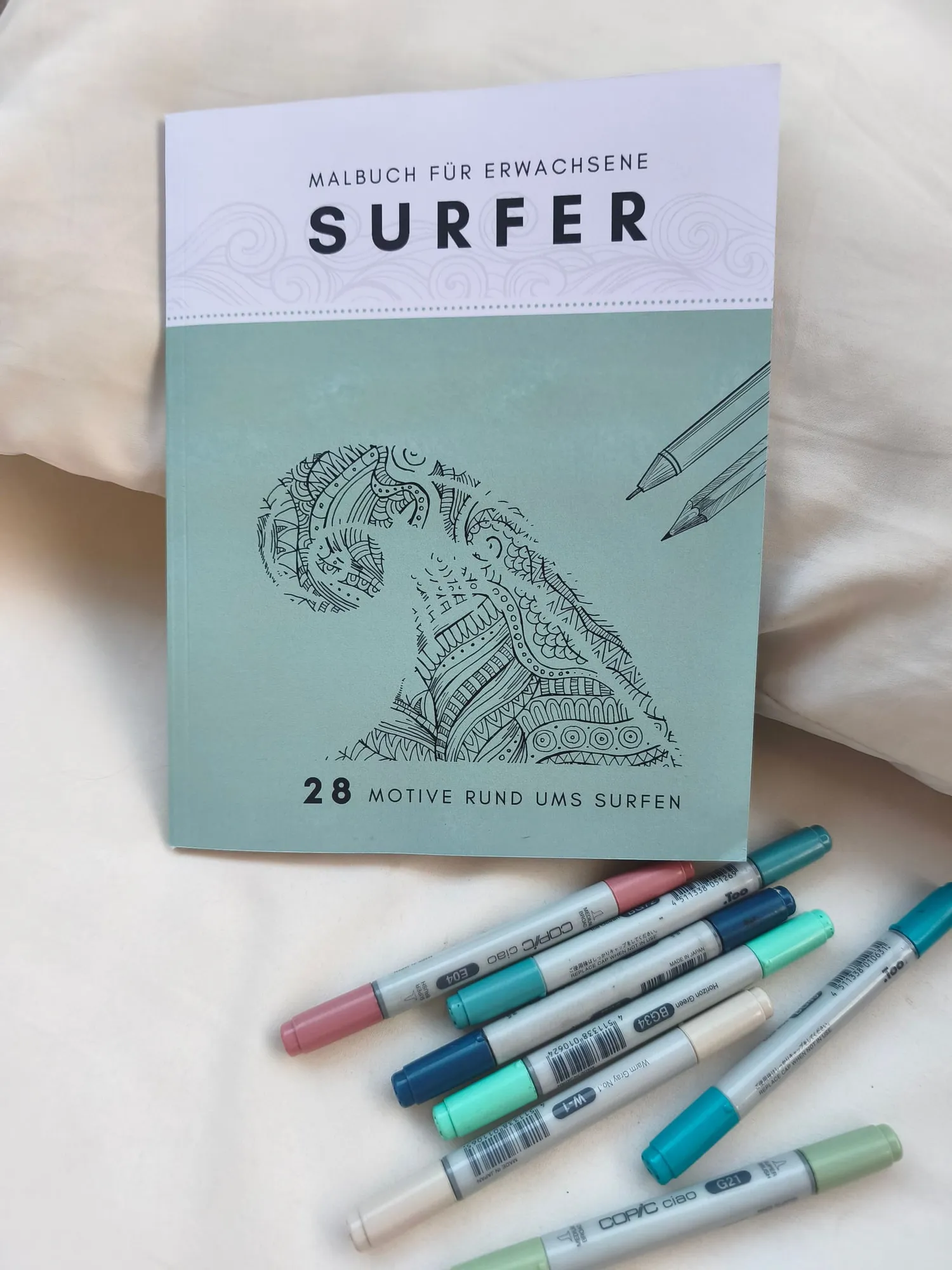 Das geschlossene Malbuch liegt auf heller Bettwäsche, darunter fächern sich sieben Buntstiftmarker auf. Auf dem Umschlag steht „Malbuch für Erwachsene – SURFER“ über einer gezeichneten Welle, darunter „28 Motive rund ums Surfen“.
