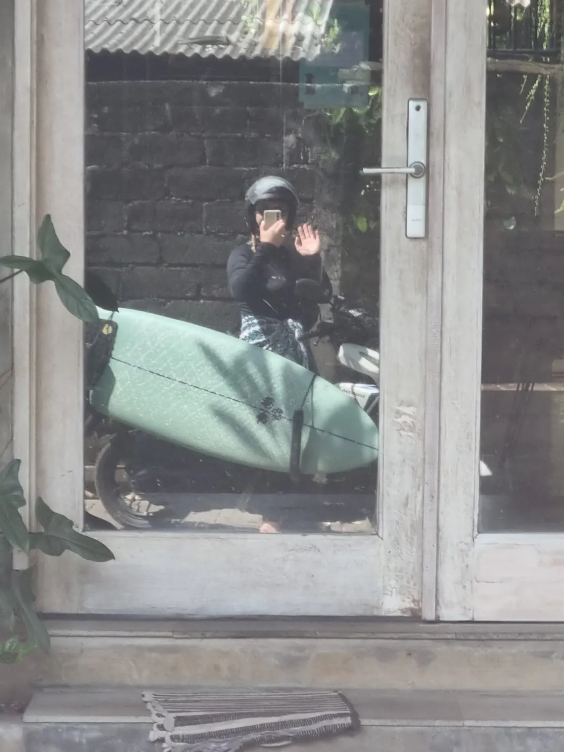 Pauline als Spiegelung in einer Glastür, winkend, mit dem Handy in der Hand; vor ihr ein mintgrünes Surfbrett auf dem Roller