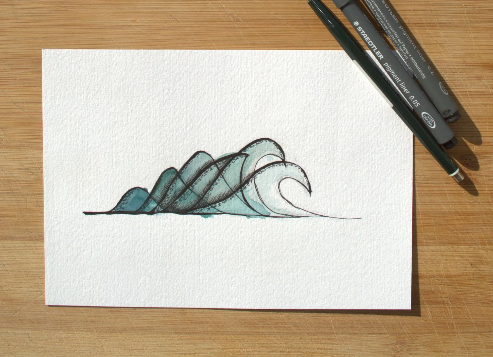 Die gemalte Welle in Türkis auf Aquarellpapier, daneben zwei schwarze Fineliner auf einem Holztisch.